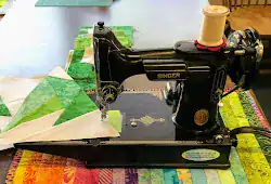 A vintage black Singer sewing machine sits on a table, stitching a colorful green and white quilt, its timeworn charm echoing memories preserved in the Feb 1 - 11 QHL Archives.