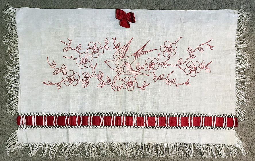 Redwork Pillow Topper A white fringed cloth features red embroidered flowers and a bird in flight, accented by a red ribbon bow and a band of red and white ribbon with lace trim—perfect for those interested in Dating Antique or Vintage Quilts.