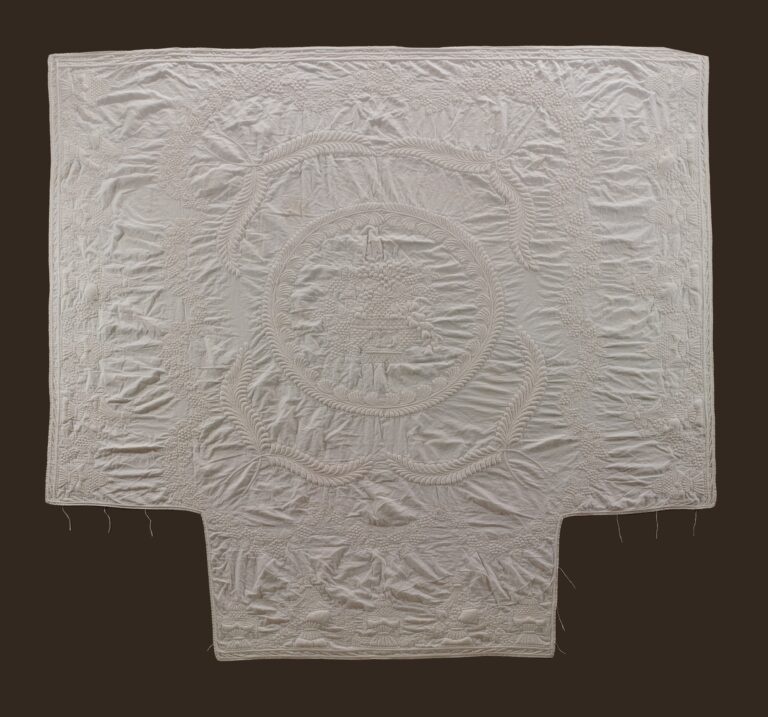 A white quilted textile with intricate stitched patterns, including a central circular motif surrounded by ornate designs. The fabric is rectangular with a notch at the bottom, displayed against a dark background.