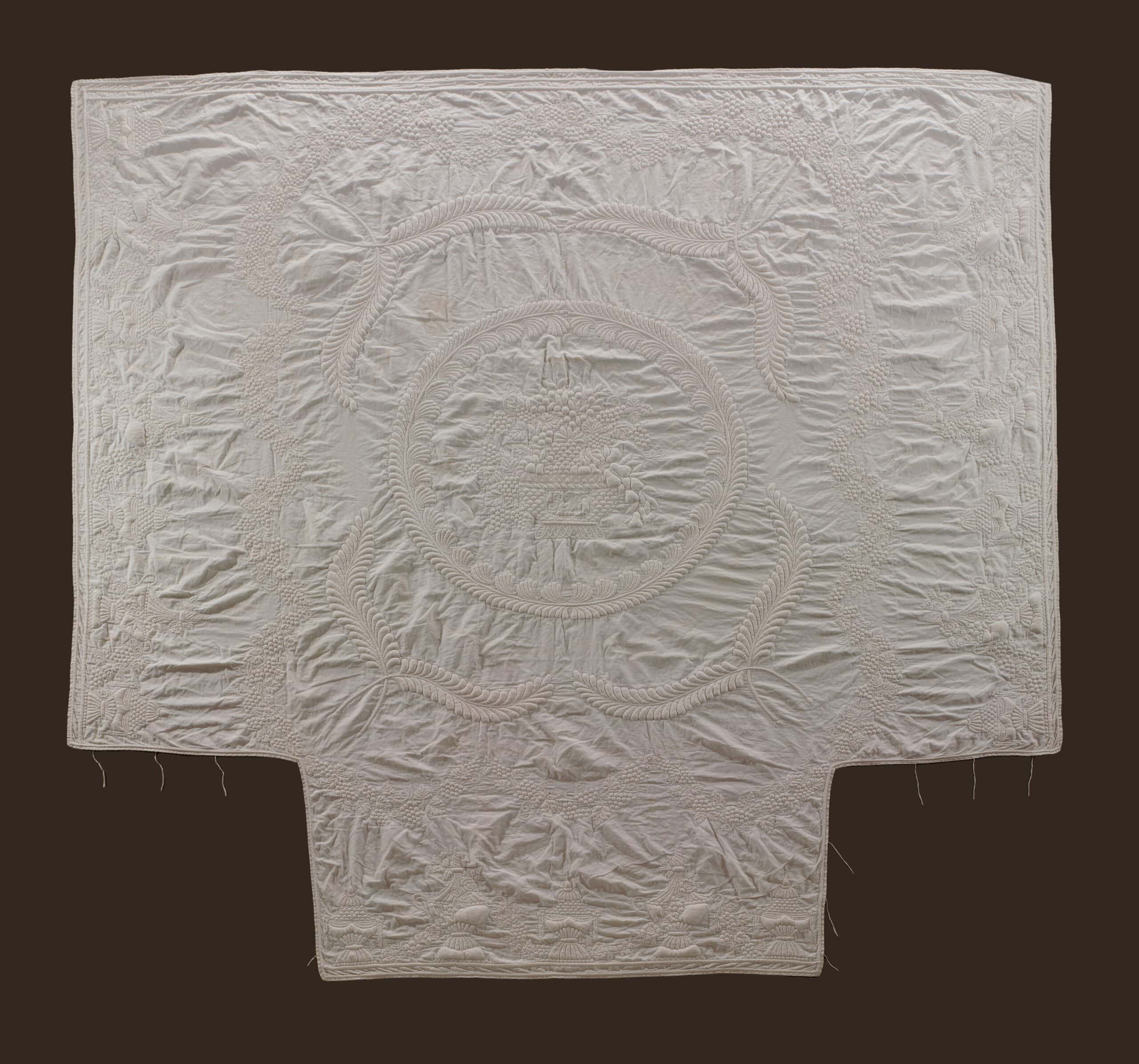 A white quilted textile with intricate stitched patterns, including a central circular motif surrounded by ornate designs. The fabric is rectangular with a notch at the bottom, displayed against a dark background.