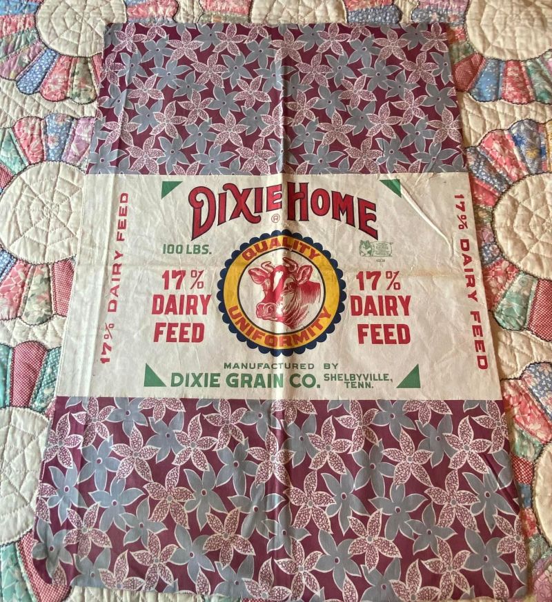 Dixie-Home-Dairy-Feed-Feedsack A vintage Dixie Home dairy feed sack with red and gray floral patterns, 17% Dairy Feed text, and a circular logo from Dixie Grain Co., Shelbyville, TN, displayed on a quilted background.