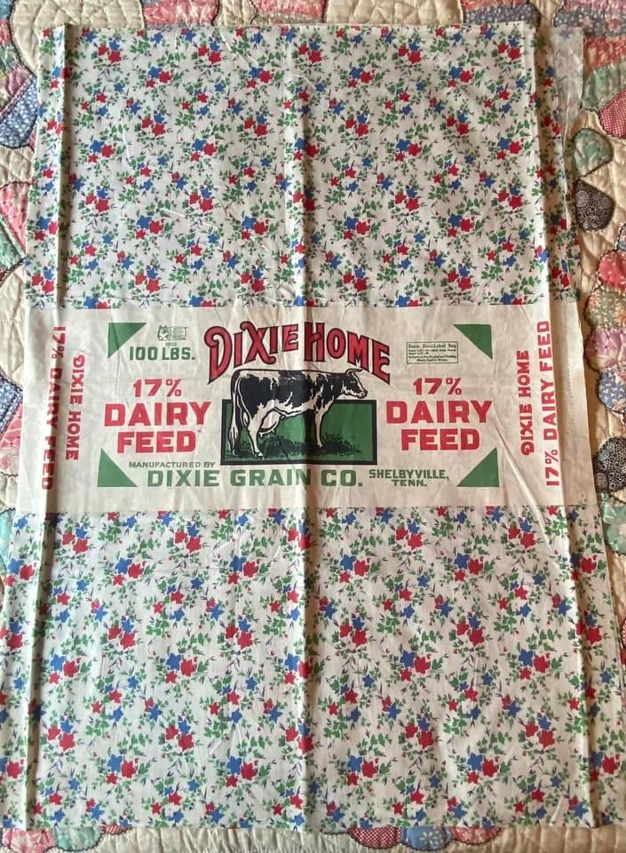 Dixie-Home-Feedsack A vintage floral feed sack with a central label reading Dixie Home 17% Dairy Feed, featuring a cow illustration and red, green, and blue flower patterns around the edges on a quilted background.