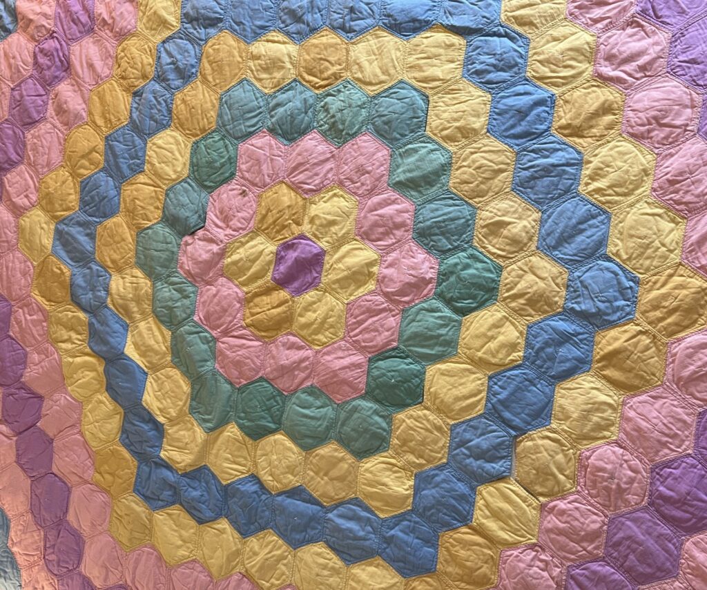 A quilt with a pattern of concentric hexagonal rings in pastel colors, including pink, yellow, green, and blue. The fabric appears slightly wrinkled, and each hexagon is outlined with stitching.