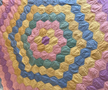 A quilt with a pattern of concentric hexagonal rings in pastel colors, including pink, yellow, green, and blue. The fabric appears slightly wrinkled, and each hexagon is outlined with stitching.