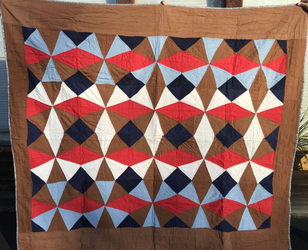 Graphic-Quilt A handmade quilt featuring a geometric pattern with triangles in brown, red, blue, and white, bordered by a solid brown edge. Two hands are visible holding the quilt at the top corners.