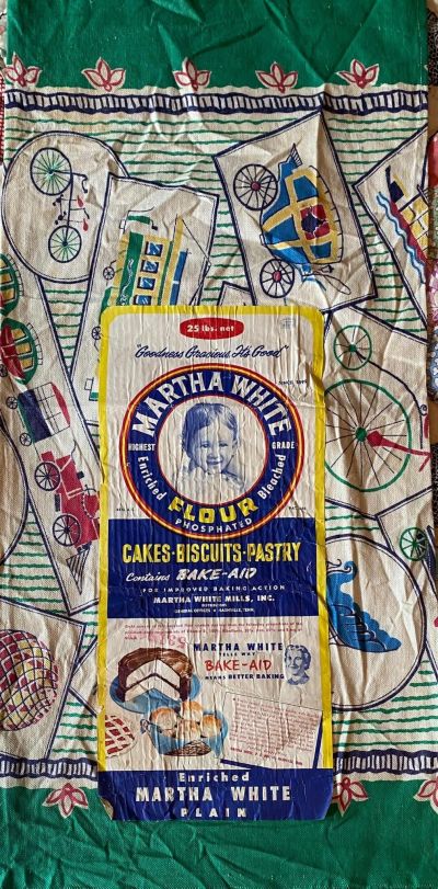 Martha-White-Flour-Feedsack A vintage Martha White flour sack sits on colorful fabric decorated with bicycles, wheels, and geometric designs. The sack has a blue and yellow label with bakery goods images and a portrait of a woman.