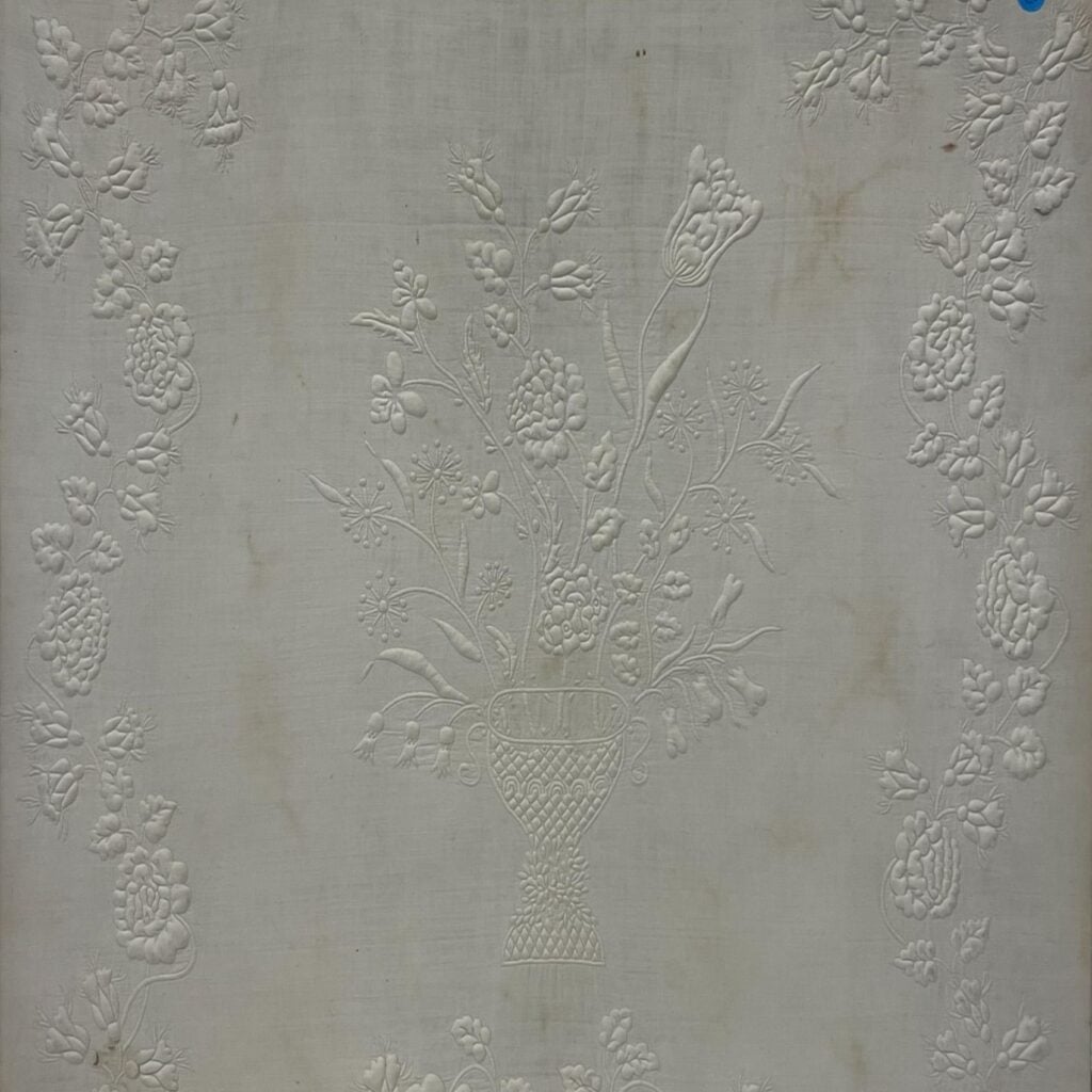 A white fabric with an embossed floral pattern, featuring a central basket filled with flowers and surrounded by decorative flower clusters along the borders.
