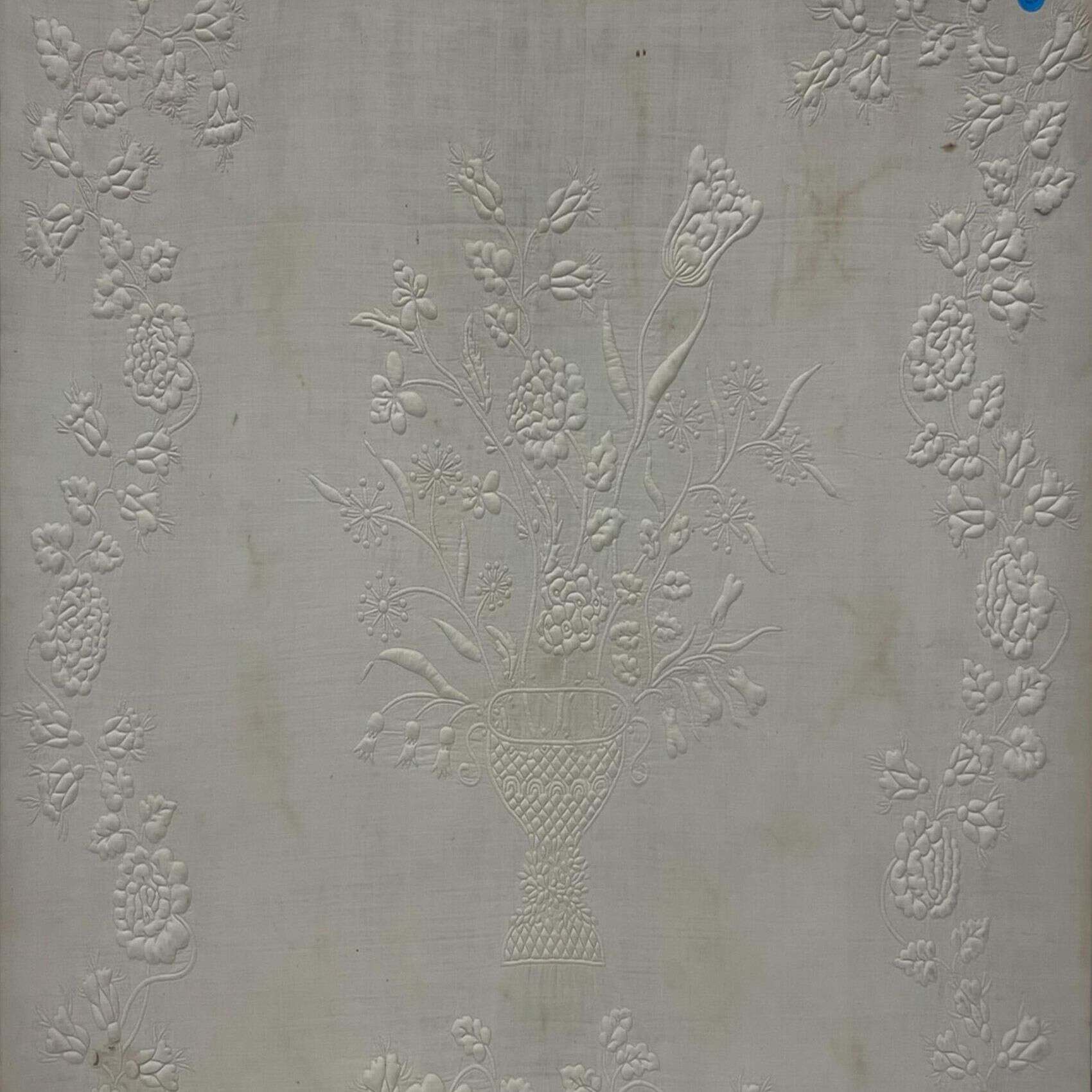 A white fabric with an embossed floral pattern, featuring a central basket filled with flowers and surrounded by decorative flower clusters along the borders.