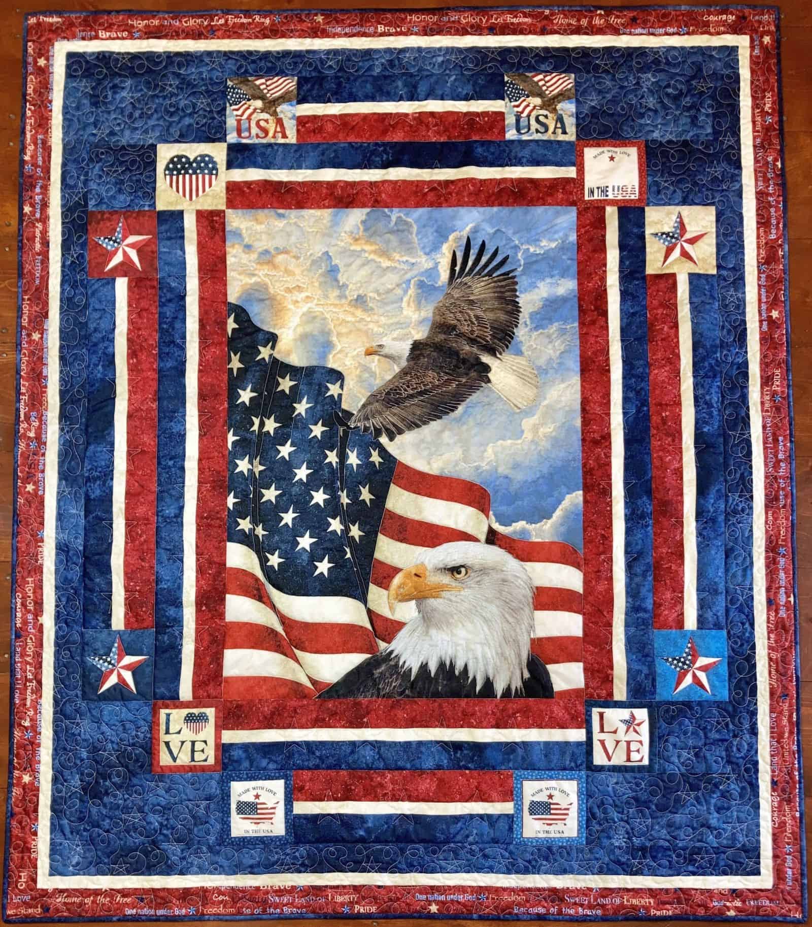 With-Love-Hilda-Panel-Quilt A patriotic quilt features a bald eagle, the American flag, stars, and USA text, accented with red, white, and blue borders. The word “LOVE” and other patriotic symbols decorate the corners.