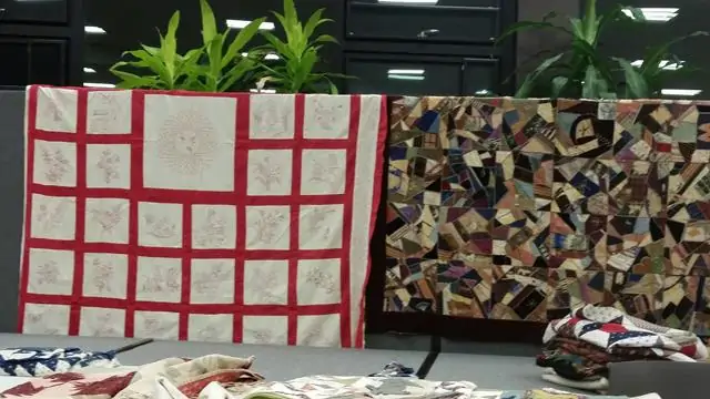 Two colorful patchwork quilts are displayed side by side on a gray surface, with green plants and office cabinets in the background. One quilt has red and white squares, the other features a random pattern of multicolored pieces.