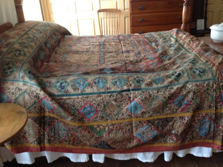chintz-quilt A large bed with a colorful patchwork quilt—reflecting Quilt History - Our Quilting Heritage—features diamond and floral patterns in blue, red, and tan. The cozy room has wooden furniture, a chair, and sunlight streaming through the window.