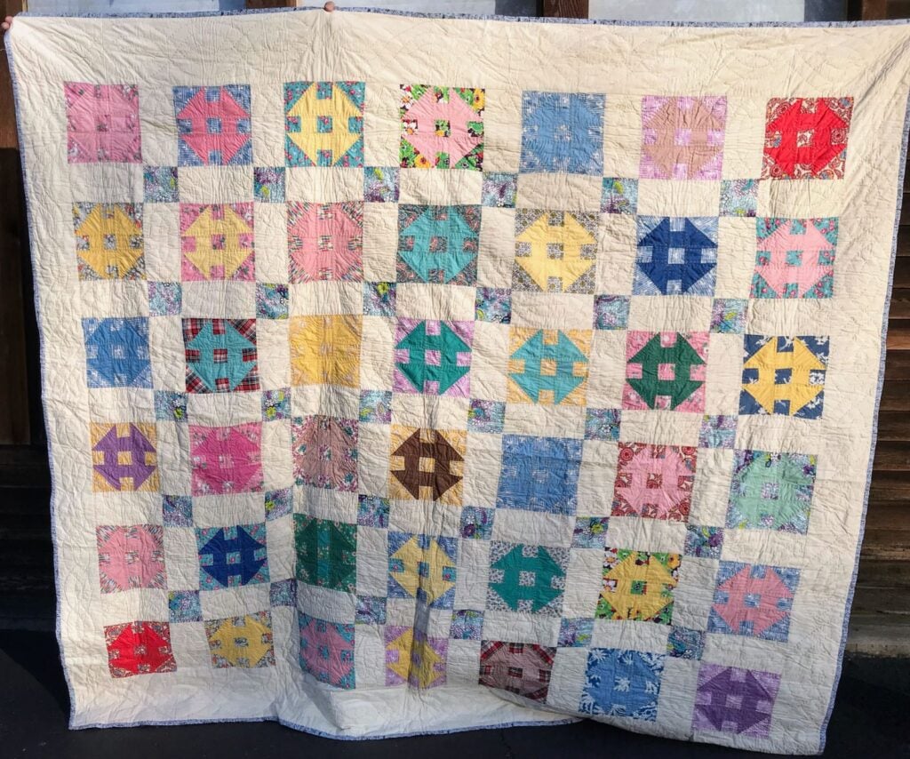 A handmade quilt with a grid of colorful patterned squares, each set within a cream border. The quilt features various pastel and bright fabrics, creating a patchwork effect, and is hanging outdoors in sunlight.