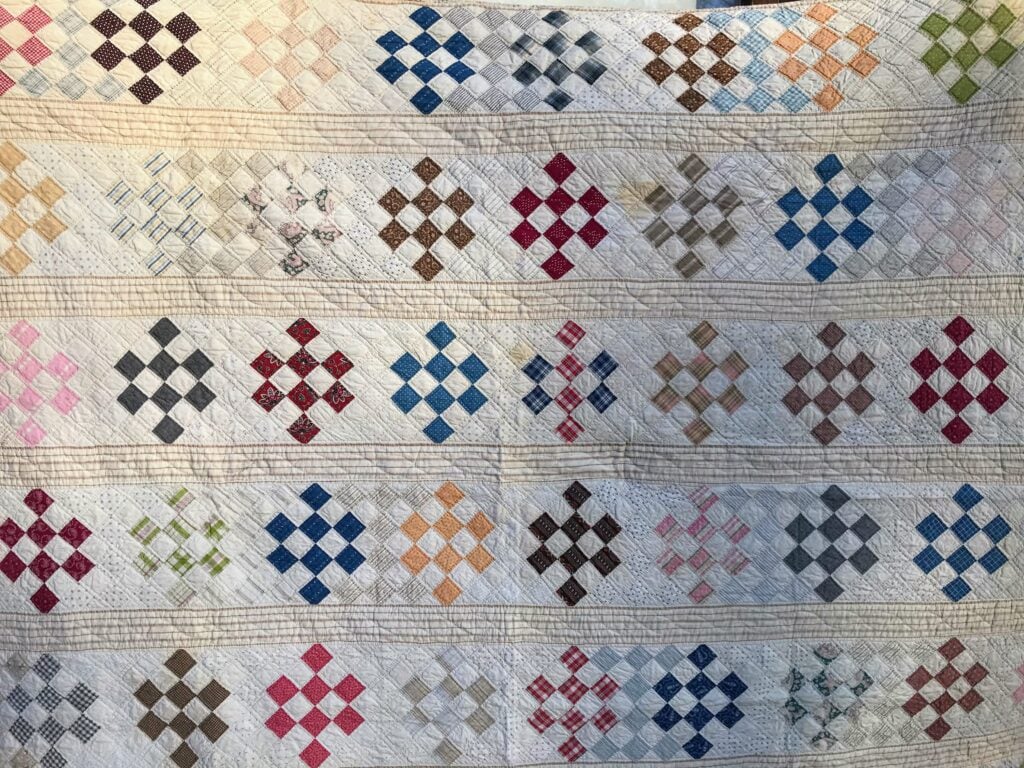 Handmade quilt featuring rows of small diamond-shaped patchwork blocks in a variety of colors and patterns on a light background, separated by thin, horizontal striped fabric bands.