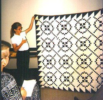 A woman stands holding up a large quilt with a geometric black and white pattern, while several people sit in the foreground observing her presentation.
