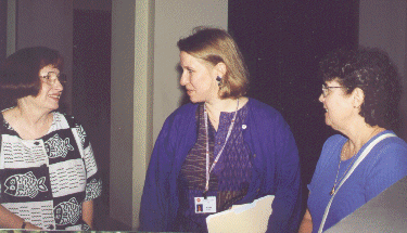 Three women are standing indoors, engaged in conversation. One woman holds folders and wears a lanyard with a badge. They all appear to be smiling or talking pleasantly.
