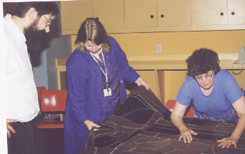 Three people examine a large, dark-colored textile laid out on a table. One person is standing at each side, while another leans over the fabric. Cabinets and red chairs are in the background.