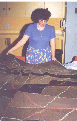 A woman with short curly hair, glasses, a blue top, and a patterned skirt is standing indoors, holding and spreading out a large patchwork quilt with diagonal lines and various shades of brown.