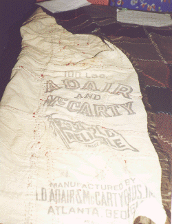 A close-up view of a quilt, showing a faded fabric label that reads L.D. ADAIR AND McCARTY and ATLANTA, GEORGIA. The quilt appears old and hand-stitched, with visible seams and stitching.