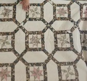 A hand rests on a quilt featuring a repeating pattern of eight-pointed star blocks within octagonal frames, using muted tones of cream, pink, green, and brown floral fabrics.