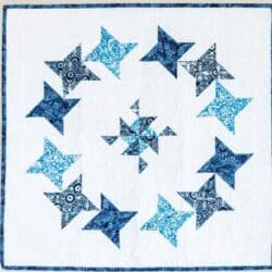A quilt featuring a white background with a circular pattern of blue and light blue star shapes, surrounding a smaller cluster of blue stars in the center. The quilt has a matching blue border.