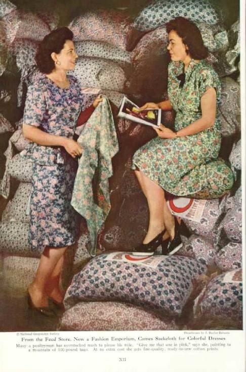 Two women wearing floral dresses sit and stand among large patterned feed sacks. One holds fabric and the other holds an open magazine as they talk, both surrounded by stacks of printed sacks.