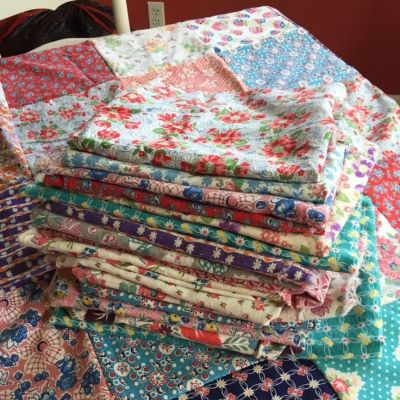 A stack of colorful, patterned fabric pieces is neatly folded on top of a patchwork quilt, with various floral and geometric designs in bright, cheerful colors.