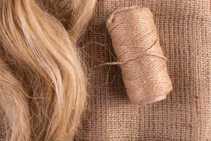 A spool of coarse, light brown twine sits on textured burlap fabric next to a bundle of blonde, wavy fibers, both displaying a natural, rustic appearance.