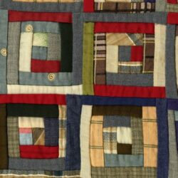 Close-up of a patchwork quilt featuring square, log cabin-style patterns made from assorted fabric strips in various colors and textures, including red, blue, green, beige, and plaid.