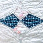A close-up of a quilt shows a diamond pattern with two blue fabric pieces featuring small white and red designs, bordered by white fabric with light pink floral accents and visible stitching.