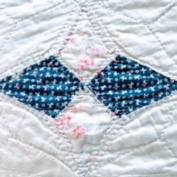 A close-up of a quilt shows a diamond pattern with two blue fabric pieces featuring small white and red designs, bordered by white fabric with light pink floral accents and visible stitching.