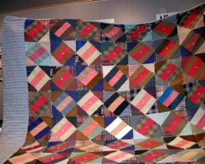A large, colorful patchwork quilt featuring a variety of patterned and solid fabric squares arranged diagonally, displayed against a wall in an indoor setting. The edges and corners have blue and striped sections.