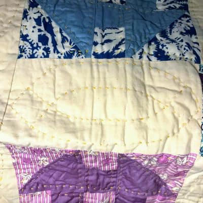 A close-up of a quilt featuring blue, white, and purple patterned fabrics stitched together, with visible yellow and white hand-stitched quilting lines forming various geometric shapes.