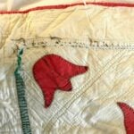 A close-up of a vintage quilt with hand-stitched red flower appliqué and green stem on white fabric. The edges are worn, and some stitching appears loose, highlighting the quilts age and handmade quality.