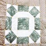 rolling-stone-quilt-block A close-up of a quilt block featuring green and white patterned fabric, with yellow knotted ties at each corner. The design includes squares and rectangles arranged in a geometric pattern.