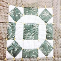 rolling-stone-quilt-block A close-up of a quilt block featuring green and white patterned fabric, with yellow knotted ties at each corner. The design includes squares and rectangles arranged in a geometric pattern.