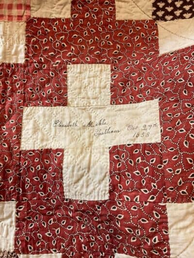 signature-block-1853 A section of a quilt features a red and white cross pattern. In the white center, “Elizabeth Michela, Chatham, Oct 27th 1858” is handwritten in cursive—a detail often used in Dating Antique or Vintage Quilts with paisley and geometric designs.