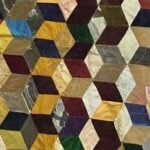 A colorful patchwork quilt features a 3D cube pattern created with various fabric pieces in shades of yellow, red, blue, green, brown, and cream. The geometric design gives a sense of depth and texture.