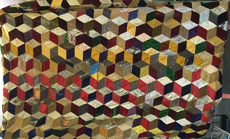 A colorful quilt made of small fabric pieces arranged in a 3D cube pattern, using shades of yellow, red, blue, green, brown, and beige in a geometric, eye-catching design.