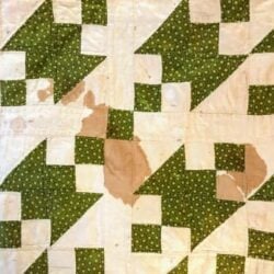 A quilt with a white and green geometric pattern has several noticeable brownish stains on its surface, mainly in the center of the quilt tiles. Ideal for those interested in cleaning antique and vintage quilts.