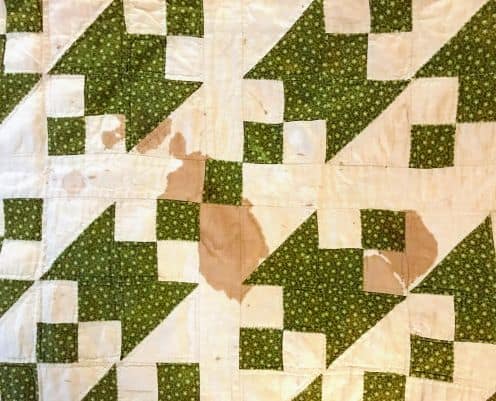 A quilt with a white and green geometric pattern has several noticeable brownish stains on its surface, mainly in the center of the quilt tiles. Ideal for those interested in cleaning antique and vintage quilts.