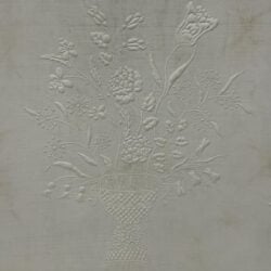 Embossed floral design on fabric, featuring a basket filled with detailed flowers and stems, all in white, creating a textured, raised pattern against a plain background.
