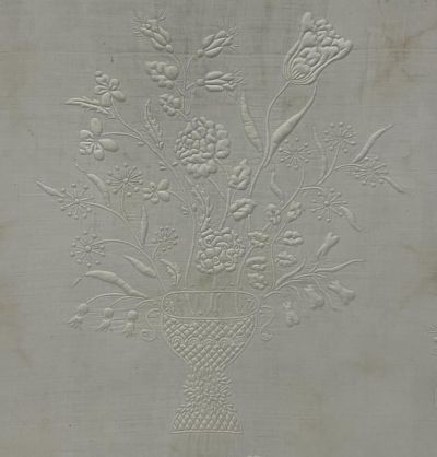 Embossed floral design on fabric, featuring a basket filled with detailed flowers and stems, all in white, creating a textured, raised pattern against a plain background.