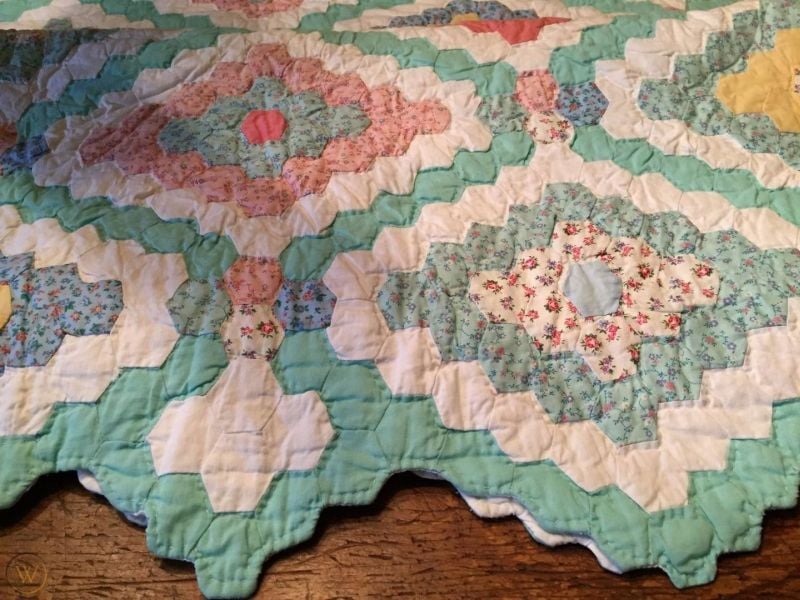 A close-up of a handmade quilt with a scalloped edge, featuring pastel colors and floral patterns arranged in a geometric, zigzag design. The quilt is laid on a wooden surface.