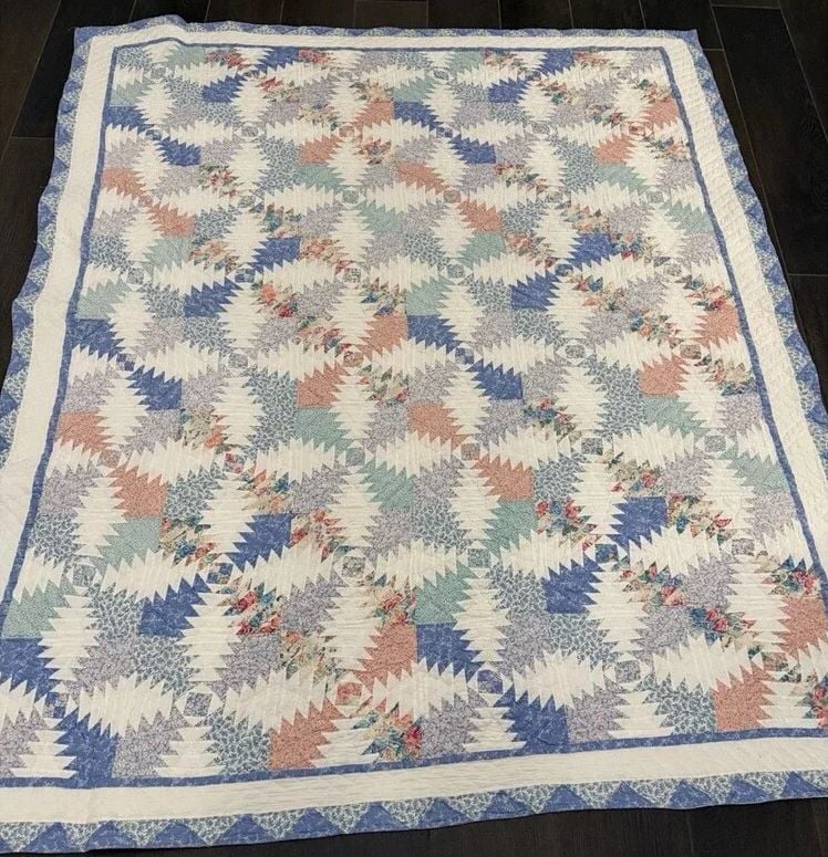 Handmade quilt with a geometric pine tree or zig-zag pattern in pastel shades of blue, pink, lavender, and white, bordered by blue and white floral and striped designs, laid flat on a dark surface.