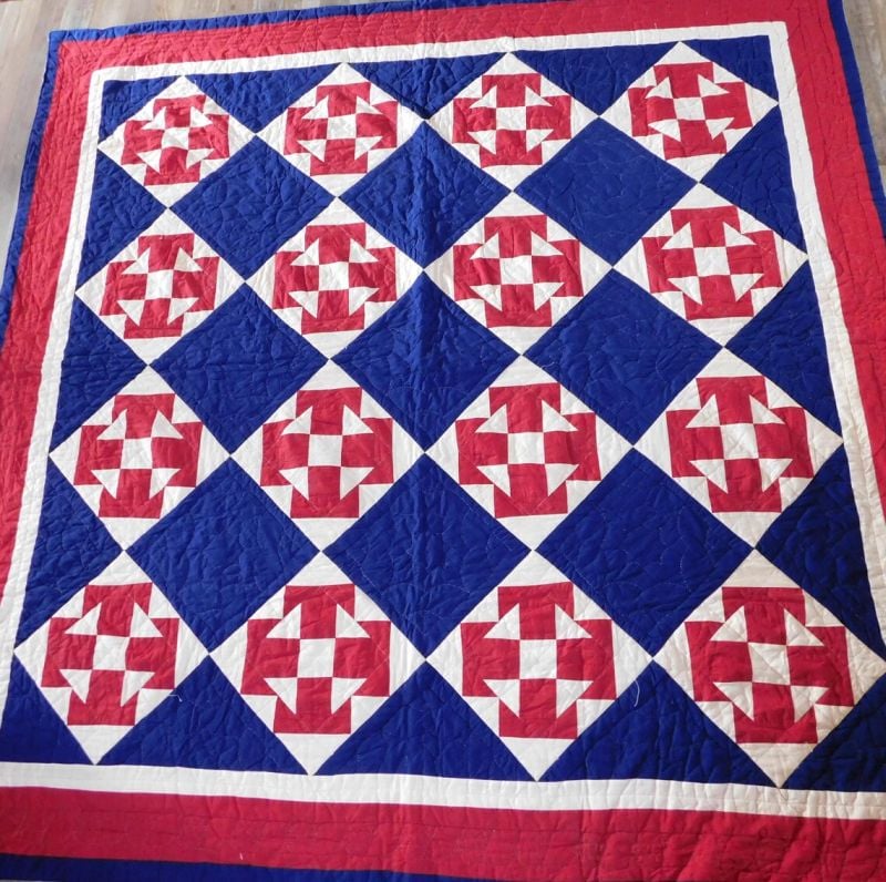 A handmade quilt featuring a repeating geometric pattern of red, white, and blue shapes arranged in diamond blocks, bordered by red, white, and blue strips. The quilt has a traditional, patriotic design.