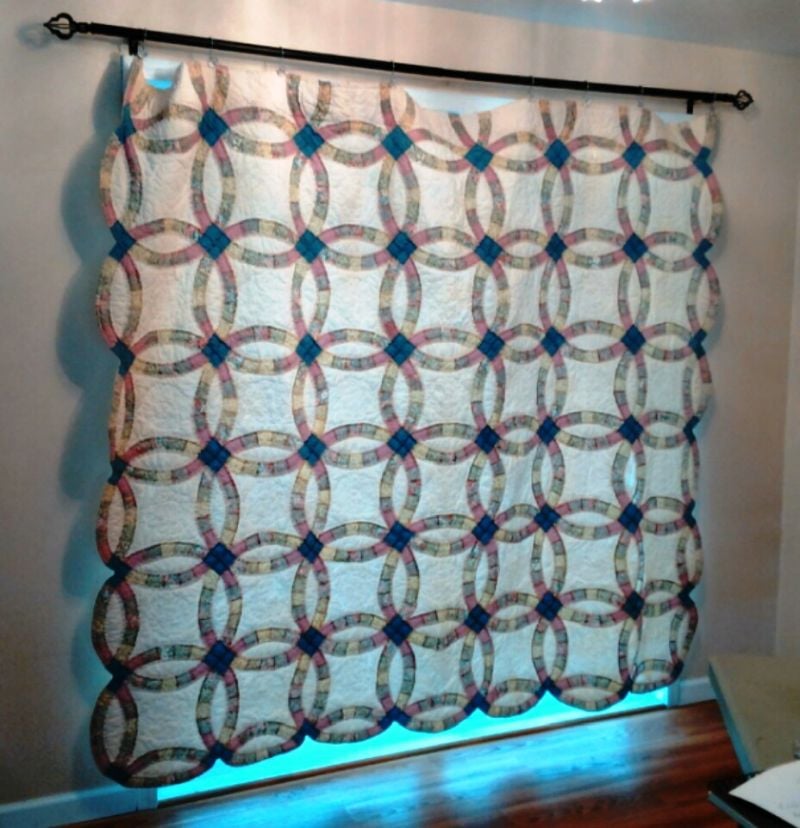 A large quilt with an interlocking double wedding ring pattern in blue, pink, and cream tones hangs on a rod against a white wall, with sunlight peeking from beneath it onto a wooden floor.