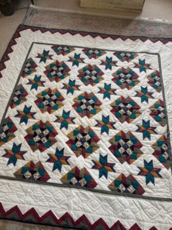 A handmade quilt featuring a repeated star pattern in teal, burgundy, gold, and white, bordered with a zigzag maroon edge, is spread out on a rug. The quilt has intricate geometric and floral designs.