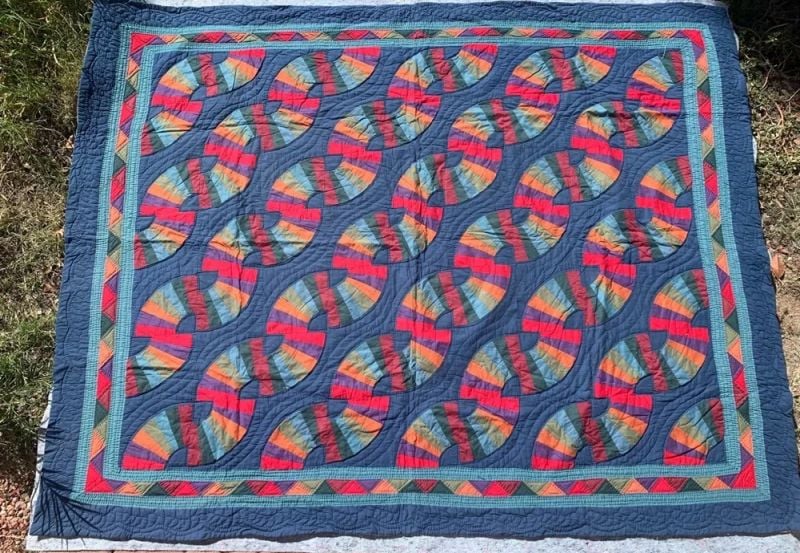 A colorful quilt laid on grass features a repeating fan pattern with red, blue, green, and gold sections, bordered by geometric designs on a dark blue background.