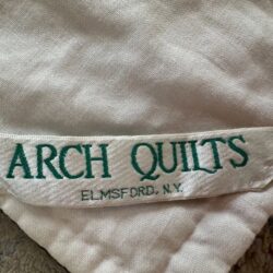 A fabric label sewn onto a quilt reads ARCH QUILTS in green text, with ELMSFORD, N.Y. printed below in smaller letters. The label is attached to a cream-colored quilt.