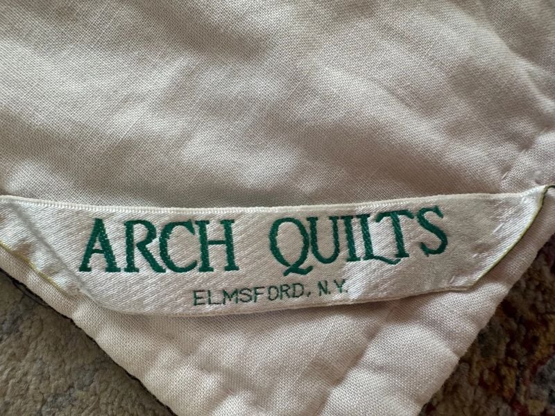 A fabric label sewn onto a quilt reads ARCH QUILTS in green text, with ELMSFORD, N.Y. printed below in smaller letters. The label is attached to a cream-colored quilt.
